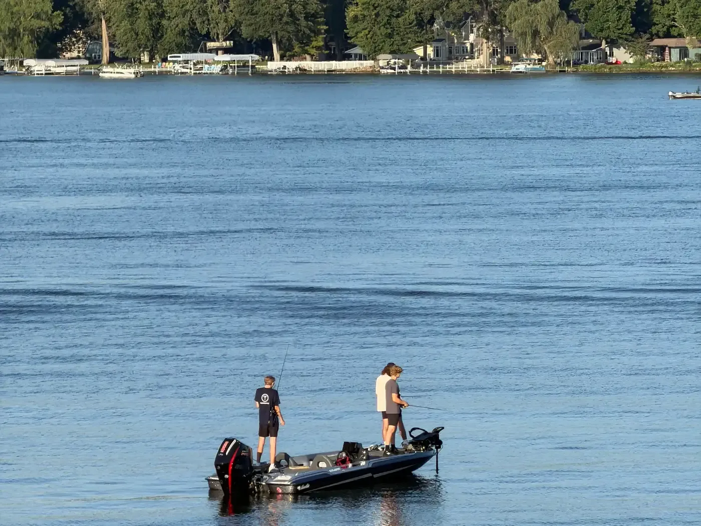 Three anglers fishing from a bass boat on calm water at Winona Lake, with docks and trees along the far shore