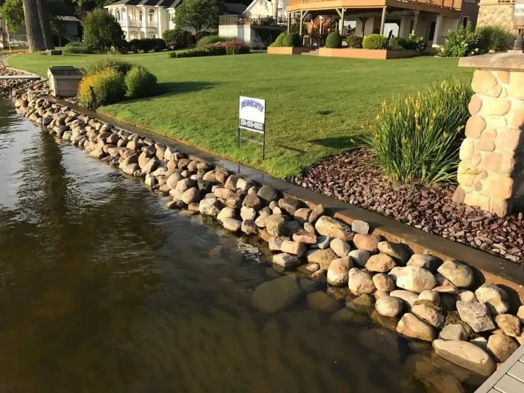 A glacial stone shoreline edge below a lawn and landscaped beds at a lakefront home