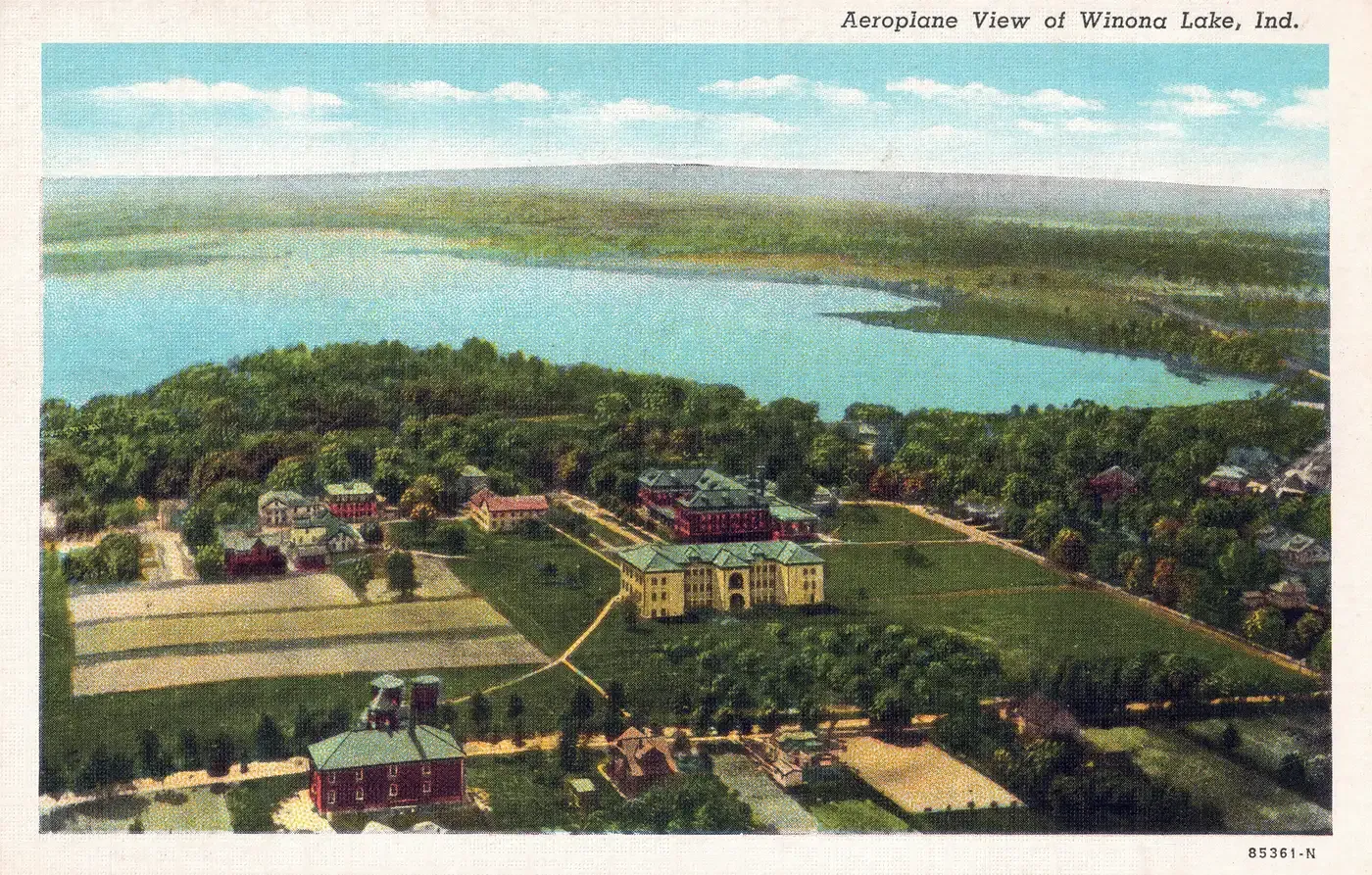 An aerial view over the Winona Lake assembly grounds, with a large cream building with a green roof and a red brick building on open lawns, cottages among trees, and the whole lake and its far shore beyond, titled Aeroplane View of Winona Lake, Ind.