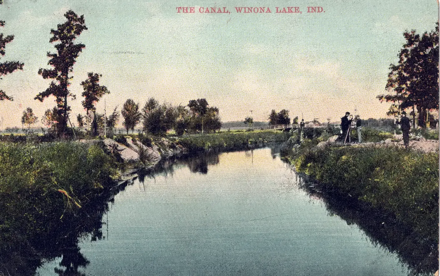 A straight canal between grassy banks and tall trees, with a footbridge and a few people at the far end and open farmland beyond, titled The Canal, Winona Lake, Ind.