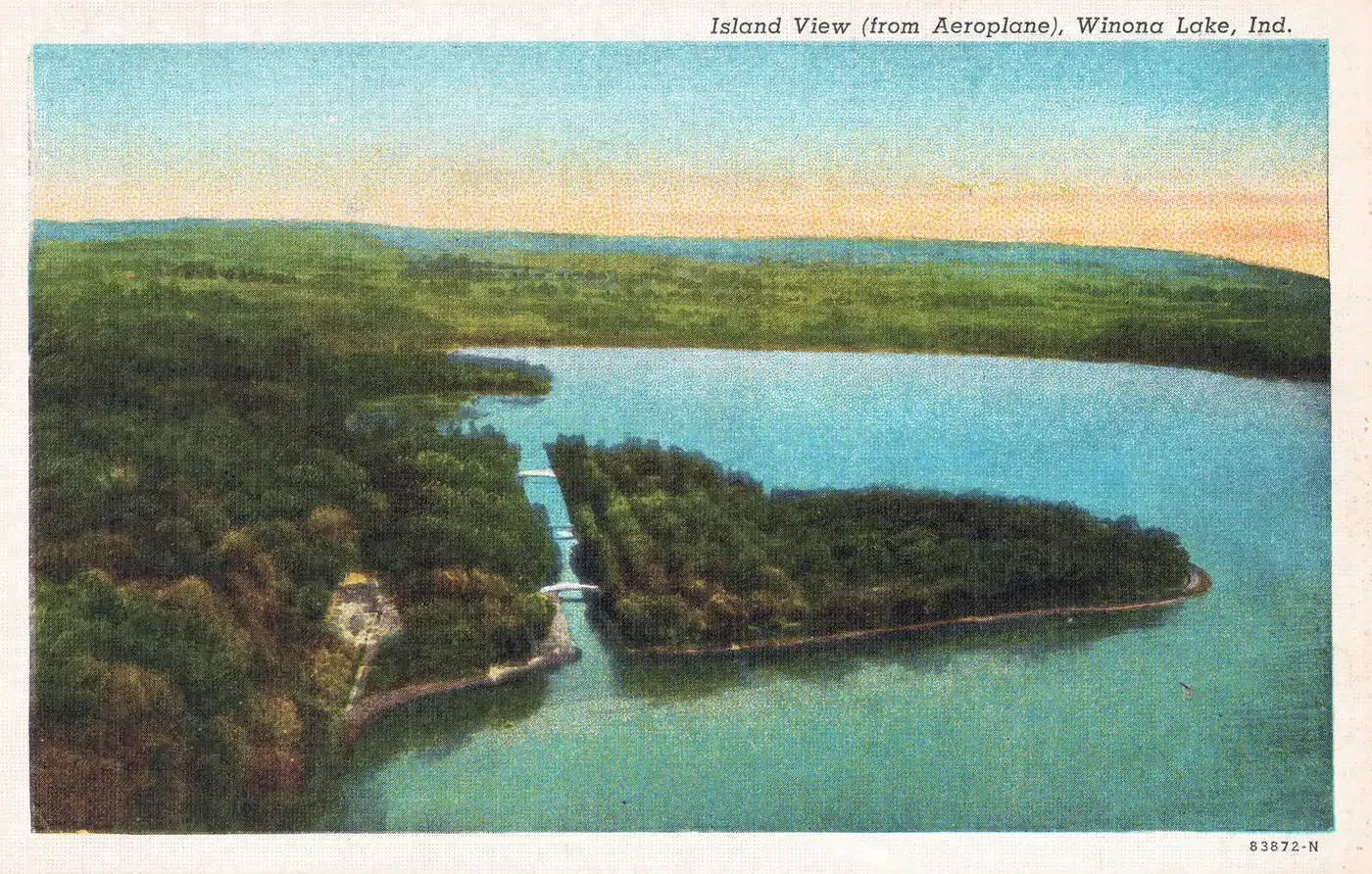 An aerial view of a wooded island separated from the wooded shore by a straight canal with two bridges, with open farmland and the far shore beyond, titled Island View (from Aeroplane), Winona Lake, Ind.