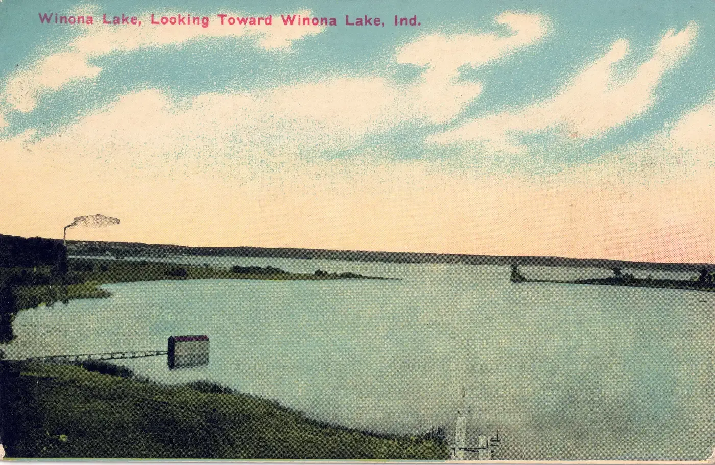 A view across calm water toward a low far shore with a long pier and a small pavilion on it and a smokestack at the left, under a blue sky with white clouds, titled Winona Lake, Looking Toward Winona Lake, Ind.