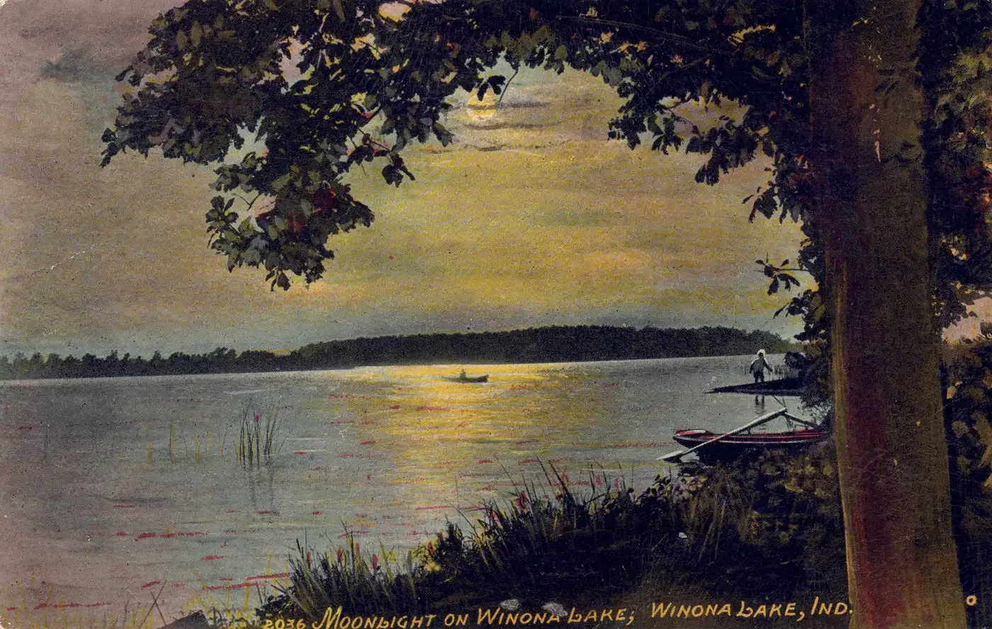 A shoreline at night under a large tree, with a rowboat pulled up on the bank, a figure at the water's edge, a small boat out on the moonlit water, and a dark far shore, titled Moonlight on Winona Lake, Winona Lake, Ind.