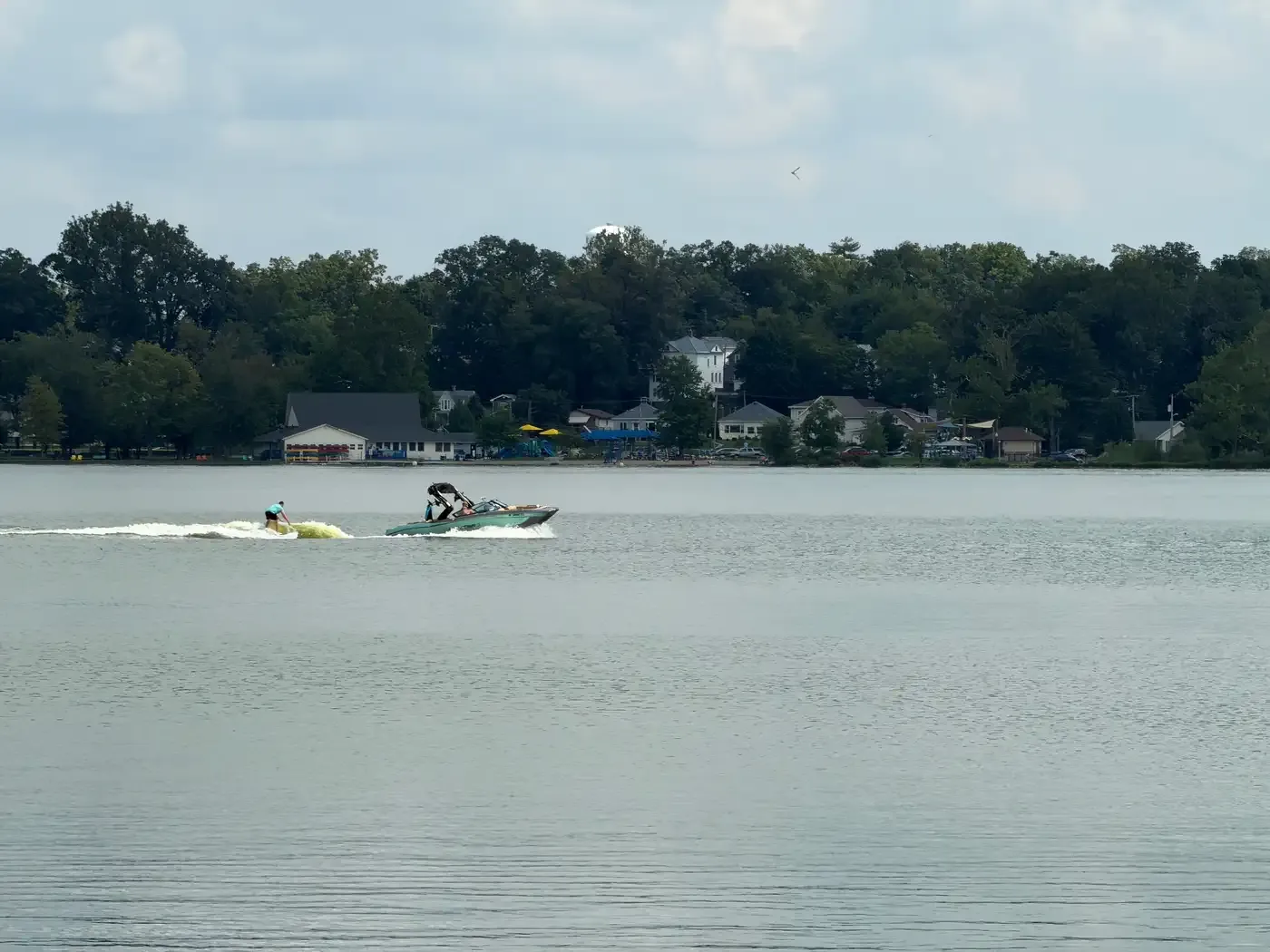 A wake boat towing a surfer mid-lake on Winona Lake, with the Limitless Park shoreline in the distance